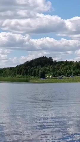 The video shows a wide shot of a lake with a forested hill in the background. The sky is blue with scattered white clouds. On the shore of the lake, there are several tents and a few vehicles, suggesting a campsite. Some blue boats are also visible on the water near the shore. The water is rippled, indicating a light breeze. The overall scene depicts a tranquil natural setting with signs of human activity.