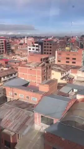 The video shows an aerial view of a city. The buildings are predominantly brick, with some having tin roofs. There are water tanks visible on the roofs of some buildings. There is a cell tower situated among the buildings. The sky is blue with scattered clouds.