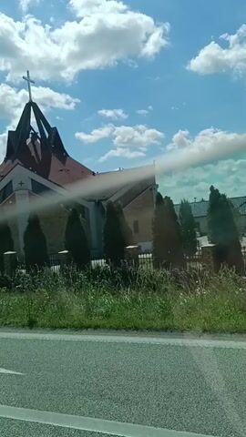 This media file is a short video clip showing a modern-looking church building from the perspective of a moving vehicle. The church has a distinctive, angular roofline with a prominent cross at its apex. The building appears to be constructed with brick and has large windows. In front of the church, there is a strip of green grass and some bushes, with a fence visible behind them. The foreground of the video shows a portion of a road and a white line marking the edge of the road. A horizontal bar, likely a part of the vehicle's interior, is visible across the middle of the frame, partially obscuring the view of the church.

The scene is set outdoors during the daytime, under a bright blue sky with fluffy white clouds. The sunlight suggests it is a pleasant day, possibly late morning or afternoon. The time stamp "00:00" to "00:04" indicates the clip is very short, about 4 seconds in duration. The perspective of looking out from a vehicle implies the church is being viewed as one passes by, possibly on a journey. There are no people visible in the clip. The overall impression is a quick glimpse of a religious building in a semi-rural or suburban setting, viewed from the road. The location context provided is Lipno, Poland. There is no visible text within the media file itself.