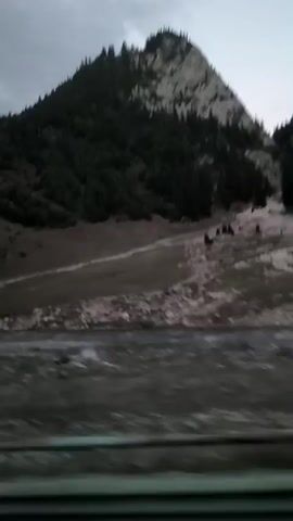 The video pans across a mountainous landscape with a cloudy sky. The mountains are covered with evergreen trees, and there are rocky outcrops visible. A body of water, possibly a river or stream, flows in the foreground. The terrain appears rugged and undeveloped, with some patches of grass and scattered rocks. The overall impression is one of natural, untamed wilderness.