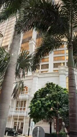 The video shows the exterior of The Ritz-Carlton Galaxy hotel. The camera angle begins behind a palm tree and pans up and to the right, revealing the hotel's entrance and facade. The building features a white base with gold accents and a tall, glass-clad tower. The entrance has a golden awning. A black car is parked near the entrance. A "Galaxy" banner is visible on a pole. There are manicured green bushes and trees around the hotel. The sky is overcast.
