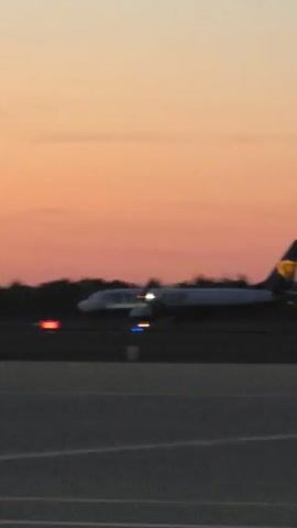 The video captures an airport tarmac at dusk, with the sky painted in hues of orange and yellow from the setting sun. Silhouetted against the colorful sky are trees and airport buildings. The primary focus is a Ryanair airplane, seen from the side as it taxis or prepares for takeoff. Lights on the runway and the aircraft itself are visible, illuminating the scene. The ground is a dark tarmac with white markings. In the foreground, a blurred segment of a person's head suggests the viewpoint of someone observing the scene. The overall mood is tranquil and atmospheric, characteristic of an airport at the end of the day. The visible text "RYANAIR" is clearly printed on the side of the airplane.