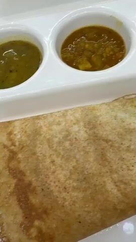 The video shows a rectangular white serving tray with a South Indian meal. The meal includes a folded dosa, a thin crepe made from fermented batter, and a set of three condiments served in small white bowls. The condiments appear to be a yellow sambar with visible vegetables, a green chutney, and what looks like a deep-fried vada. The tray is set on a white surface, and a metal knife and fork are placed alongside the dosa. Another similar tray with the same condiments is visible in the background.