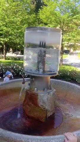 The video shows a close-up of a water fountain outdoors. The fountain has a large glass cylinder at the top filled with clear water and ice. A metal pipe extends from the bottom of the cylinder into the water. A tap on the side of the metal pipe is open, and a stream of water is flowing out. The water falls into a large, circular basin that is filled with dark brown water. The basin appears to be made of concrete and has a rough texture. The base of the fountain is made of rough, brown and grey rocks. In the background, there are green trees and a small child wearing a blue hat and white shirt. The lighting suggests it is daytime.