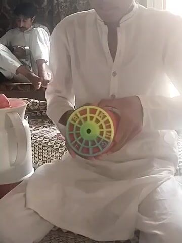 The video clip features two men relaxing indoors. In the foreground, a man, dressed in a white traditional shalwar kameez, sits on a patterned rug. He is holding and examining a bright red kite reel with a colorful (yellow, green, and blue) spoked hub. To his left, a white and red thermal pot or kettle is visible on the rug.

In the background, another man, also wearing a white shalwar kameez, sits on a raised platform or low sofa, looking down at an object in his hands. His bare feet are visible.

The setting appears to be a casual living area in a home. Bright natural light streams in from the right side of the frame, suggesting it is daytime. An audible Islamic call to prayer (Azaan) plays in the background throughout the clip, indicating a specific prayer time. The overall atmosphere is calm and relaxed, with both individuals engaged in quiet activities within what appears to be a typical residence in Dera Chahal, Pakistan. There is no visible text in the frame.