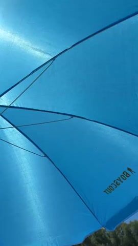 The video shows an upward view from under a blue beach umbrella. The sun is shining brightly through the fabric of the umbrella, creating a light blue, textured effect. The umbrella's metal spokes are visible, as is the white central pole. The bottom edge of the umbrella is fringed and hangs over a grassy area with some trees and a body of water in the distance. There are also some pieces of wood in the foreground on the ground. The scene appears to be outdoors on a sunny day.