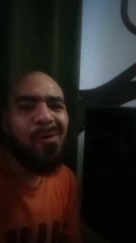 A man with a shaved head, dark eyebrows, and a short beard is looking directly at the camera. He is wearing an orange t-shirt. He initially smiles widely and makes a hand gesture resembling a small heart shape. His expression then becomes more serious, with a slightly furrowed brow. The background includes dark green curtains on the left and a dark object, possibly a television or speaker, on the right. The lighting is dim, casting shadows on his face and making the background partially obscure.