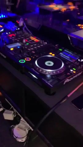 The media file shows a close-up of a DJ setup in a dimly lit establishment, likely a club or bar, with purple and orange lighting. The main subject is a Pioneer DJ controller, specifically a CDJ-2000 Nexus, with its illuminated jog wheel and numerous buttons and faders. A person's hands are visible operating the mixer, adjusting knobs and faders, indicating a DJ is actively playing music. The background is blurred but shows tables and chairs, suggesting a seating area for patrons. The overall atmosphere appears to be nighttime, given the artificial lighting and darkness. There is text visible on the DJ equipment, including "Pioneer" and "CDJ-2000 Nexus". Another piece of text on a white coiled cable on the floor reads "HIGH QUALITY PROFESSIONAL CABLE". The scene is static, with the focus on the DJ equipment and the actions of the DJ. The location context of Tiraspol, Moldova is provided but not visually discernible from the media itself.