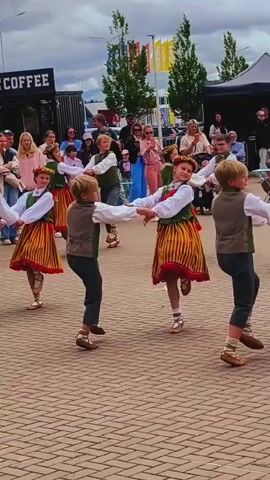 In a medium shot, a group of children perform a folk dance outdoors on a cobblestone surface. Six children, three boys and three girls, are visible. The girls wear long-sleeved white blouses, dark green vests, and long, multi-colored striped skirts with red trim at the bottom. They also wear floral wreaths on their heads. The boys wear white shirts, dark green vests, dark pants, and light-colored socks with dark straps over them. All children wear similar light-colored shoes. They dance in pairs, holding hands and spinning, with some children facing the camera and others with their backs to it. The background shows a crowd of adults and children watching the performance, some standing, some seated. To the left, a dark food truck with the words "ENJOY YOUR COFFEE" in white letters is visible. The sky is overcast.