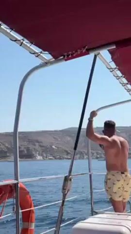 The video shows the view from a boat on a body of water, most likely the ocean. The boat has a red canopy and a white safety rail. A man stands holding onto the rail wearing shorts and no shirt. There is a life preserver on the boat. The landscape in the background consists of dark, rocky hills with what appear to be some boats anchored offshore. The sky is a clear, bright blue.