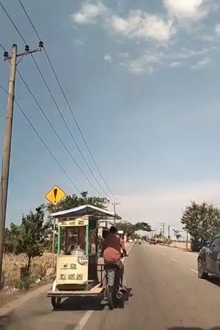 A camera follows a man riding a motorcycle with a cart attached to it on a paved road. The cart appears to be a food cart, potentially selling bakso as suggested by the audio.  The road is straight, and there are power lines overhead. To the left is some dry vegetation and a yellow caution sign. A car drives in the opposite direction in the right lane. The sky is blue with scattered white clouds. The man is wearing a red or orange shirt. The video is shot from the perspective of someone driving behind the motorcycle.