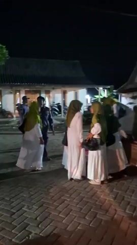 A group of women, most of whom are wearing hijabs, are gathered around the back of a red pickup truck at night. Some women are standing on the ground, while others are in the truck bed. One woman in the truck bed is reaching out her hand, possibly to help someone down. The truck is parked on a brick-paved area, and there are buildings in the background. A motorcycle is visible in the foreground, blurred by motion. The lighting suggests an outdoor event or gathering.