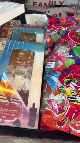 The video shows an assortment of sweets in boxes and packages on a counter. There are at least six different kinds of packaged candies. Many of the boxes are branded with El Shanawany. The packages contain nougat, Turkish delight, and various other sweet treats. The background reveals shelves with more items, and part of a chair. The speaker says "Oh, beautiful" in Russian.