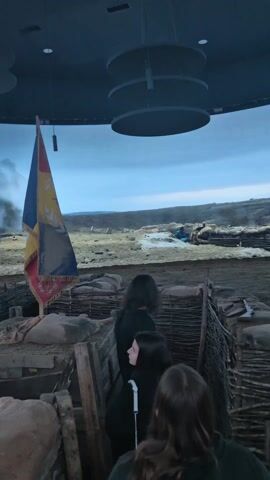 The video shows people inside a simulated trench overlooking a battlefield scene. The trenches are constructed with sandbags and wooden fences. The battlefield, depicted on a curved screen, shows soldiers in blue uniforms behind similar trenches, with smoke rising in the background. The sky is overcast. The viewer appears to be within a museum exhibit, given the immersive and theatrical nature of the setting.