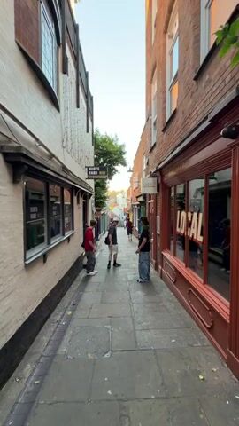 The video shows a narrow alleyway in Nottingham, England, identified by a street sign reading "Hurts Yard NG1". The alley is paved with stone and lined with buildings, some of which house small shops. Several people are present. Initially, three young men are standing near the alley entrance. As the camera moves forward, other individuals can be seen further down the alley, walking or standing. One man with dreadlocks is seen running. The shops are decorated with various signs and artwork, adding color to the scene. The overall impression is of a bustling, vibrant urban space.