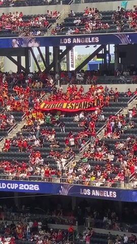 The video shows a crowded stadium filled with spectators wearing red and yellow, the colors of the Spanish flag. The crowd is predominantly viewed from above and behind, with a large banner displayed across the stands that reads "FURIA ESPAÑOLA." Above the banner, the word "LOS ANGELES" is visible on a stadium structure. Below the banner, another sign reads "WORLD CUP 2026 LOS ANGELES." People are seated in tiered stadium seating, with many standing and some holding flags. The overall atmosphere appears to be that of a sporting event, likely a soccer match, given the context of the World Cup and the nationalistic display. The lighting suggests it is daytime.