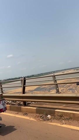 The video shows a view from a vehicle moving across a bridge.  A metal safety railing lines the edge of the bridge, and the road surface is visible. Beyond the railing is a wide riverbed, mostly exposed sediment and sand. A few buildings are visible in the distance, along the riverbank. The sky is partly cloudy and bright.