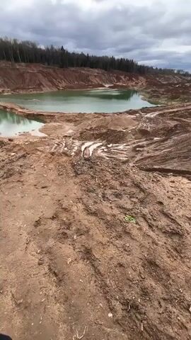 The video shows an overcast day at a quarry with a small, green-tinted lake. Initially, the camera pans over the muddy, tire-tracked ground and the lake. A man in a black hooded jacket appears, looking downcast. The camera then shifts to a dirt road lined with trees. Lastly, the man and a woman wearing a beige jacket and grey beanie appear together, standing against the backdrop of the quarry lake. The woman is smiling at the man, while he looks away.