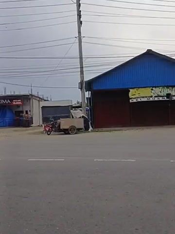 The video shows a street view of various small shops and stalls. The scene is shot from a vehicle moving along a paved road. On the left side of the road, there are several buildings, some with awnings and signs. One building has a sign that says "PRIMA" and displays prices. Another building appears to be a car wash with a black tarp covering part of it. There is also a small, rustic structure with a thatched roof. A motorcycle is parked in front of one of the stalls. Further down the street, there are more shops, including one with a blue and yellow facade and another with a red and green facade. A tall cell tower is visible in the background. The sky is overcast.
