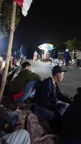 The video shows a nighttime scene with a group of people gathered along the side of a paved road. In the foreground, several individuals are sitting on blankets on the ground, facing away from the camera. One person in a black baseball cap is looking to the right. Further down the road, there is a small food stall with a colorful umbrella and bright lights. More people are standing and sitting around the stall and along the roadside. A white pole with a red and white flag is visible on the left side of the frame. The environment appears to be rural, with trees and vegetation visible in the background. The overall atmosphere suggests a communal gathering or event taking place after dark.