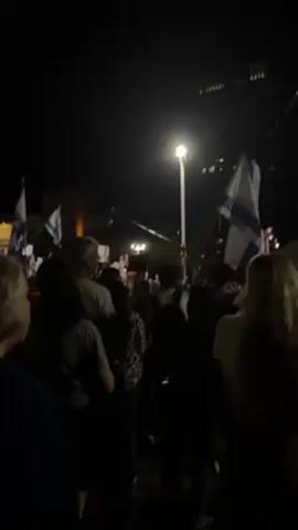 The video shows a nighttime shot of a crowd holding Israeli flags. The camera is positioned within the crowd, looking towards a slightly elevated area. The crowd is primarily composed of adults, and they seem to be focused on something in front of them. The environment is dark, but there are streetlights providing some illumination. There's a building visible in the background with lit-up windows. A few people are holding their flags high, and the overall atmosphere appears to be one of collective focus and perhaps participation in an event or gathering.