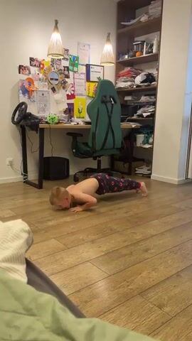 A young boy is doing push-ups on a wooden floor in a room. He is shirtless and wearing dark pants with a red and white pattern. He is in a room that appears to be a home office or study, with a desk, computer, chair, and shelves filled with books and various items. The desk has a gaming steering wheel attached to it. The wall behind the desk is decorated with pictures and certificates. Two pendant lights hang from the ceiling. The room appears to be indoors, with artificial lighting. The boy seems determined and focused on his exercise. The scene suggests a casual, domestic setting. There is no visible indication of the time of day or weather. The location context is Veliky Novgorod, Russia.