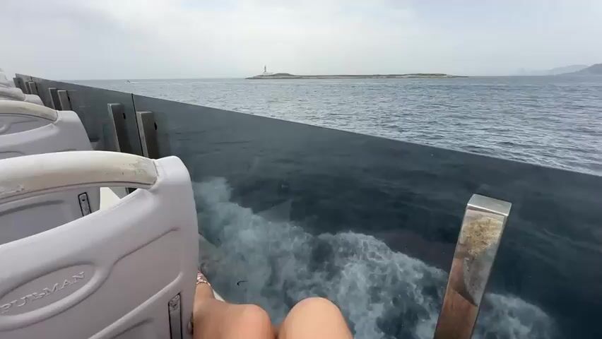 The video shows a view from a moving boat looking towards a distant island with a lighthouse. The camera is positioned behind a dark, reflective barrier, possibly a window or railing, with white seats visible in the foreground. The water is choppy, creating white foam and spray as the boat travels. The sky is overcast, with a light grey hue. The island is small and rocky, with a white building and a striped lighthouse on it. Other islands or landmasses can be seen faintly in the background. The overall scene conveys a sense of travel and exploration on the sea.