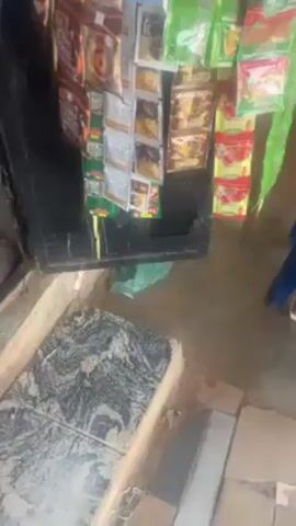 The video shows a close-up of a small retail stand. The stand is made of dark wood, and it's covered with various food items hanging in single-serving packets. The items include packets of Quaker White Oats, Chin Chin snacks, and various soup seasonings in green and red packaging. A granite-like tile is on the ground in front of the stand. A blue plastic chair leg is visible on the right side of the frame. The audio appears to capture a conversation, potentially related to a purchase or request for items.