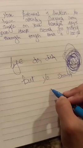 This is a close-up, overhead shot of a notebook with lined paper. In the upper half of the notebook, in blue ink, is a handwritten inspirational quote that reads: "Your potential is limitless. You have already survived every single on your heart. Get days put back. Echoes for getting through tough times is 100%". Below this quote, also in blue ink, is another handwritten phrase that appears to read: "life do, but yo should Smile". The bottom right of the frame shows a person's right hand holding a blue pen, actively writing in the notebook. The hand is positioned to the right of the second phrase, and the pen is writing the word "Smile". There is a circular doodle with scribbled lines to the right of the second phrase and above the hand. The notebook is resting on a surface that is not clearly visible. The lighting suggests an indoor environment.