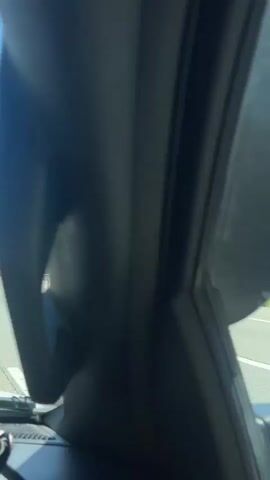 The video captures a first-person view from inside a vehicle driving on a highway during what appears to be a sunny day. The camera is held by someone in the passenger seat, with their hands and a smartphone visible in the reflection of the side mirror throughout the clip. The side mirror also shows the text "OBJECTS IN MIRROR ARE CLOSER THAN THEY APPEAR."

The vehicle is moving past a section of road with orange traffic cones lining the right side of the lane. A white guardrail separates the road from a dense forest of green trees that runs alongside the highway under a bright blue sky with a few white clouds.

As the vehicle progresses, two road work trucks become visible ahead, each with a large, orange, diamond-shaped sign on the back displaying "ROAD WORK AHEAD" in black lettering. The trucks appear to be involved in ongoing road construction or maintenance, with barriers and additional cones marking the work zone. The trees in the background are vibrant green, indicating either spring or summer. The overall scene suggests travel through a natural, possibly wooded, area that is currently experiencing road improvements.