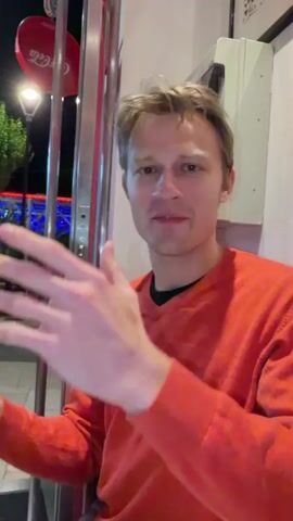 A man wearing a reddish-orange sweater is standing inside a building with a Coca-Cola sign visible outside the entrance. He's holding a pair of black glasses in his hands. He appears to be talking and gesturing enthusiastically with his hands. Outside the building, there are trees, a lamp post, and what looks like outdoor seating with lights.  The man has blond hair and is looking directly at the camera. The video is shot at night.