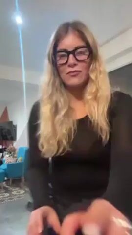A woman with long blonde hair, wearing large black-rimmed glasses, a black long-sleeved shirt, and black pants, stands in a room and moves to the rhythm of music that is playing. She is smiling and pointing with her finger while mouthing the words to the song. The room has a white ceiling, a gray floor, and a turquoise chair is visible in the background. A television is visible on a wall and art is hanging on the back wall. There is a bright light source overhead. The video is shot from a low angle.