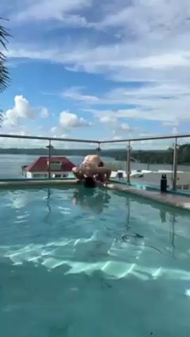 A man in red swim trunks and headphones kneels by a light blue swimming pool on a rooftop with a glass railing. He briefly gestures towards the camera, smiling, before looking away. Behind him is a view of a body of water with a red-roofed building on the nearby shore. The sky is blue with puffy white clouds. There is a black water bottle near the railing.