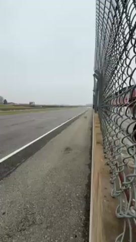 The video shows a neon green sports car speeding down a racetrack on a cloudy day. The video is shot from behind a chain-link fence along the side of the track. A man wearing a black jacket can be seen briefly in the left side of the frame next to the fence.