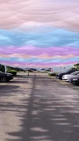 The video pans upwards to reveal an outdoor scene with a vast expanse of draped fabric creating a colorful canopy. The fabric is arranged in horizontal layers, with distinct bands of color: pale pink at the top, followed by layers of light blue, purple, and pale orange. This creates a visual effect reminiscent of a layered sky or aurora borealis. Below the fabric canopy, a paved pathway or road is visible, lined with parked cars on either side. The cars appear to be of various makes and models, mostly sedans and SUVs, parked in a lot. Further in the background, a low-rise building with an arched entrance and windows is present, with some greenery and trees surrounding it. A few people are seen walking on the pathway in the distance. The lighting suggests it is daytime, possibly late afternoon, with shadows cast on the ground by the cars and the fabric above. The overall impression is an art installation or decorative setup in an open-air parking area or public space.