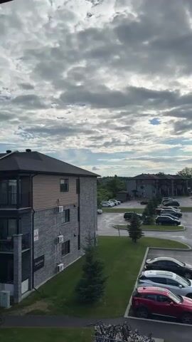 This is an aerial shot of an apartment complex parking lot. The apartment buildings are made of stone and siding, with multiple stories and peaked roofs. There are many cars parked in the lot, mostly SUVs and sedans. The parking lot is wet, suggesting it has recently rained. Several small evergreen trees are planted throughout the complex, adding a touch of nature to the urban environment.

The sky is cloudy, with patches of sunlight breaking through, indicating it is likely daytime, possibly late afternoon or early evening. The clouds are dramatic and dynamic, creating a sense of atmosphere. The scene is peaceful and quiet, with no people visible in the immediate vicinity. The overall impression is one of a typical residential neighborhood in a Canadian city. The visible text on the building reads "D'Argenteuil".