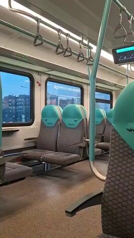 The video shows the interior of a train car. The seats are upholstered in a gray fabric with a patterned design, and the headrests are a teal color. Each headrest has a logo with a bird silhouette and the word "ИВОЛГА" printed on it. There are grab handles on the ceiling and a teal-colored pole in the aisle. Through the windows, buildings and other train cars can be seen, suggesting the train is in motion or at a station. A hand briefly enters the frame from the left side of the screen at 00:02, obscuring part of the seats. The lighting suggests it is either dusk or dawn outside.