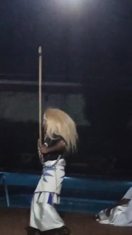 A man is dancing in front of a pool at night. He is wearing a black shirt, a white skirt with blue designs, a headband, and a long blonde wig. He is holding a long stick in his left hand and a small round object in his right hand. He is smiling and appears to be enjoying himself. The environment is dark and the lighting is poor, but a pool can be seen in the background.