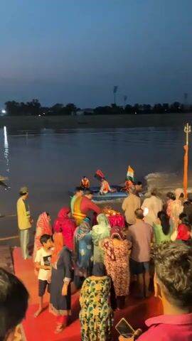 The video shows a group of people gathered at the edge of a body of water, likely a river, at dusk. There are at least two boats in the water. One is a larger, yellow passenger boat with several people on board, some wearing life jackets. The other is a smaller, blue inflatable boat with individuals wearing orange life jackets and a man in a red and gold robe. An Indian flag is attached to the side of the inflatable boat, and it is creating a wake as it moves. A group of people on the shore, including children and adults, are watching. Some are dressed in traditional Indian attire. A person in a yellow jacket is standing apart, facing the water. The overall atmosphere appears to be a cultural or religious gathering, possibly a ritual or ceremony taking place on or near the water. The lighting suggests it is either early morning or late evening.