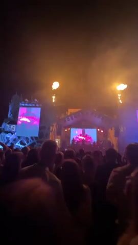 A concert is taking place at night with a large crowd gathered in front of a stage. The stage has large screens displaying visuals and is lit with colorful lights, including blue, pink, and yellow. There are also pyrotechnic effects, with flames erupting from the stage. The crowd is silhouetted against the stage lights, with some people holding up their hands. The overall atmosphere appears to be energetic and lively.