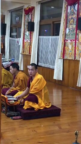 Two monks in traditional yellow and maroon robes are seated on a polished wooden floor in what appears to be a monastery or temple hall. The monk on the right sits on a maroon cushion, looking towards the camera with a neutral expression. He has an open scripture on a small wooden stand in front of him. The monk on the left is partially visible, also in robes, and is engrossed in a scripture resting on a similar wooden stand.

The background features large windows with white curtains, above which hang colorful, decorative religious banners (likely Thangkas) with intricate patterns and imagery. Black speakers are mounted on the walls above the windows. A wooden pillar is visible on the left side of the frame. A water bottle can be seen on a table in the background. The interior is well-lit, possibly by artificial light, though some soft light filters through the windows. The scene suggests a quiet moment of prayer, chanting, or scripture study. No discernible text is visible within the image.