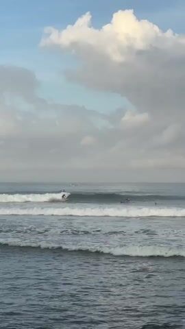 The video shows a seascape under a sky filled with dense, white and grey clouds against a backdrop of pale blue sky. In the sea, several people are visible surfing on the waves. The waves are small, white-crested, and spaced relatively close together. In the distance, small boats can be seen gently bobbing on the water. The sea itself appears dark grey, and the overall lighting of the scene suggests it is either early morning or late afternoon. The focus remains on the surfers and the waves.