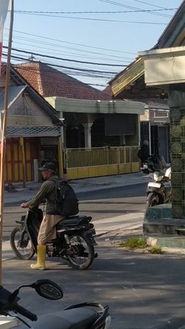 The video shows a man wearing a backpack and yellow boots sitting on a motorcycle on a street. The motorcycle is black and has a license plate. The man is looking down at the motorcycle. In the background, there are houses and other motorcycles parked on the side of the road. The sky is clear and blue. There are power lines overhead. The video is shot from a slightly elevated angle. The footage appears to be taken during the daytime.