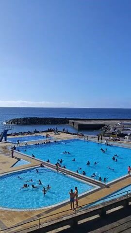 This video displays a scenic view of a seaside resort or public swimming area. In the foreground, the camera is positioned above a set of swimming pools filled with clear blue water. Several people are seen swimming, splashing, and relaxing in the pools. The pools are surrounded by a paved area with some lounge chairs visible. Beyond the pools, a rocky coastline meets the ocean, with waves gently breaking against the shore. In the distance, the vast expanse of the blue ocean stretches towards the horizon, under a clear sky. The camera pans slightly to capture more of the coastal landscape, revealing more of the resort and the natural environment. The overall impression is one of a sunny, leisurely day at a beautiful coastal destination.