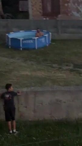 A person stands in an inflatable pool in a backyard. The pool is blue and has the Intex logo visible. The person is shirtless and appears to be moving around in the water. The backyard is covered in grass. In the background, there is a building and a fence. The scene is dimly lit, suggesting it is either dusk or night.