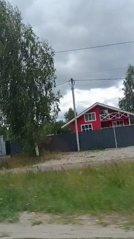 A motor vehicle is moving along a dirt road. The video shows a red house with white trim, a gray metal building, a gas station with blue and red accents, and a garden center with large green lettering that appears to say "SADOVY CENTER." Two people are walking along the side of the road. There are several trees, including pine trees and a tall leafy tree, and utility poles with wires. The sky is overcast.
