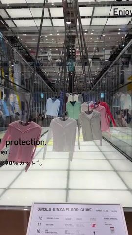 A person's face is partially visible in the foreground as they look to the right. Their right hand holds a phone and what appears to be a white paper or cloth. In the background, a store interior is visible with clothing items hanging from a ceiling structure. The clothing items, predominantly light-colored tops, are suspended by wires and arranged in rows. A digital display board is also present at the bottom of the frame, labeled "UNIQLO GINZA FLOOR GUIDE". Text overlays on the video state "UV protection." and Japanese characters. The environment appears to be well-lit with reflective surfaces.