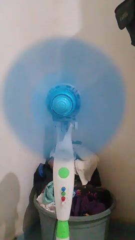 A fan is spinning. It's a standing fan with a blue fan head and a white and green base. The fan is in front of a wall, and there is a laundry basket with clothes behind it. The fan blades are blurred from spinning. The lighting is dim.