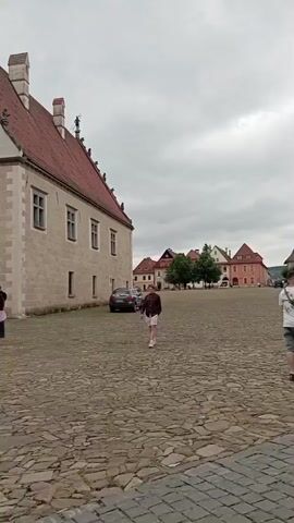 A drone shot pans across a cobblestone square in front of a pale, ornate building with a red tiled roof. A young woman in a pink top and dark leggings walks across the square away from the camera. To the right, a man wearing a brown jacket and shorts stands and faces the camera. In the background, more buildings with red tiled roofs line the square, and several cars are parked. The sky is overcast. The drone continues to pan, showing more of the square and buildings. There are other people visible in the background, some gathered in groups. The drone then focuses on a large, older building with a tall spire, suggesting it is a church or cathedral.