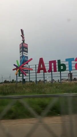 The video shows a passing view from a vehicle on a cloudy day. In the foreground, a metal fence with a diamond pattern is visible. Beyond the fence, there is a road and then a grassy area. Further in the background, a large sign with colorful letters spelling "Альтаи" is prominent. To the left of this sign is a tall vertical signpost with multiple brand logos. There are also some buildings and trees in the distance. The view continues to pan, showing more of the road and surrounding landscape, with another partial sign with red lettering reading "М-бид" appearing briefly on the right. Power lines and poles are scattered throughout the scene. The overall impression is of a commercial or retail area on the outskirts of a town or city.